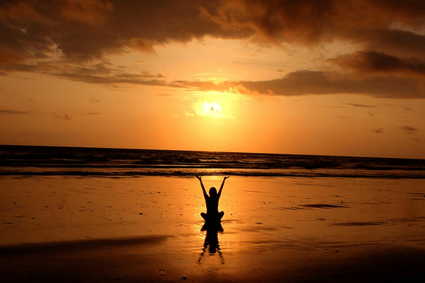 Woman meditating on a beach at sunset, arms raised in peace, surrounded by a warm and tranquil atmosphere.